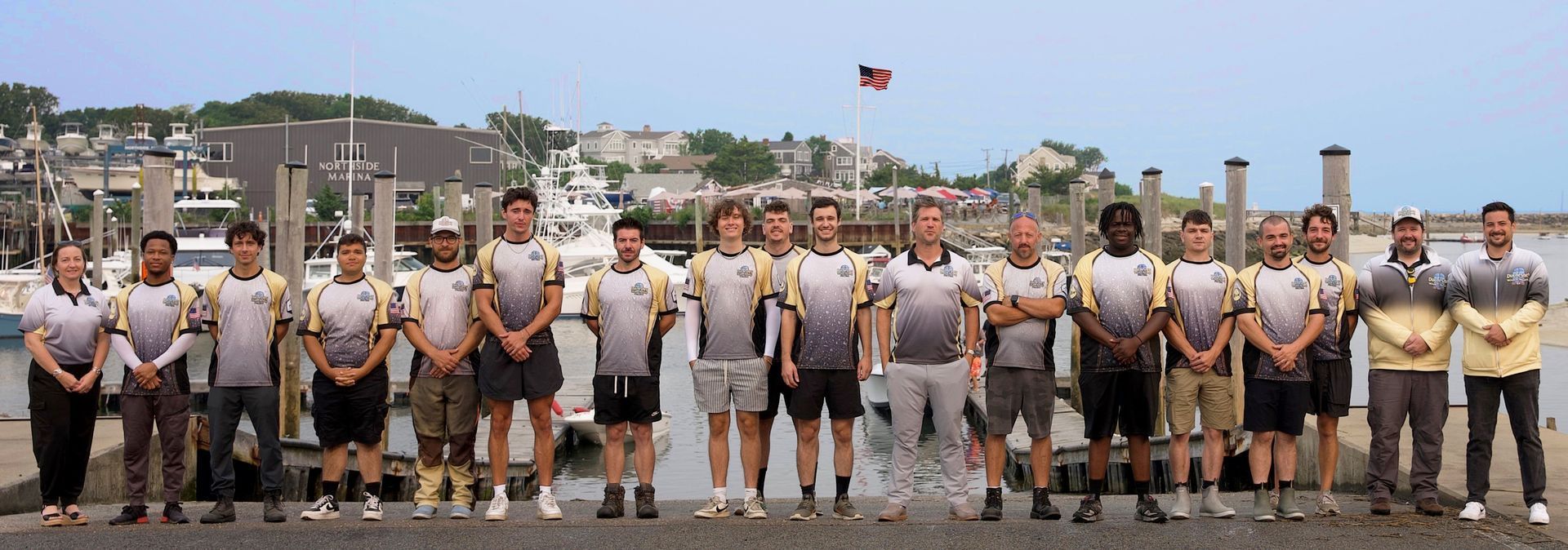 A row of uniformed people standing on a dock with boats and a seaside town in the background