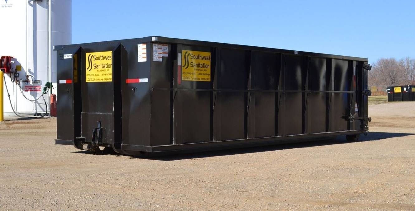 Black dumpster on gravel, outdoors on a sunny day.