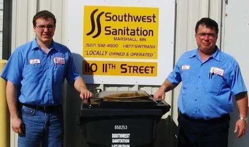 Two men in blue shirts stand by a trash bin in front of a 