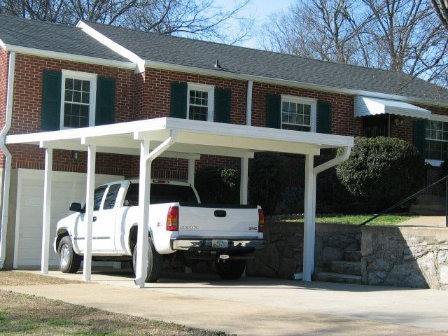 A white truck is parked under a carport in front of a brick house