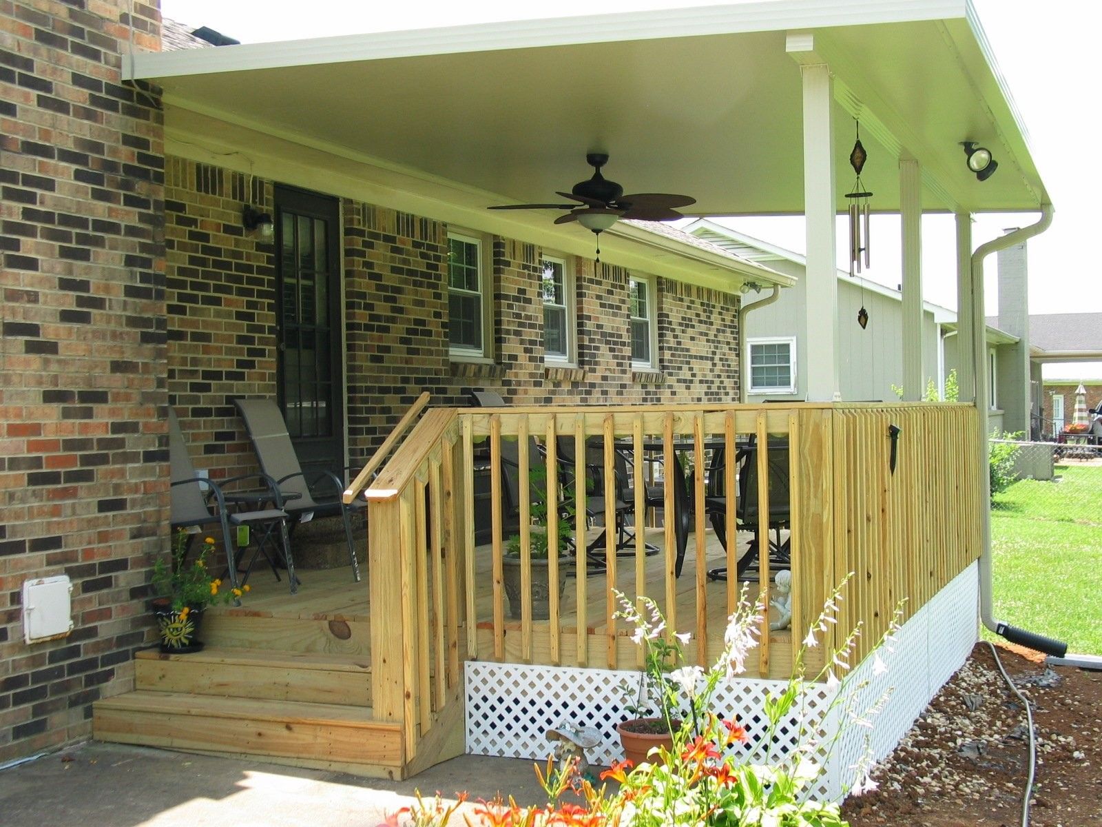 A wooden deck with a ceiling fan on top of it is in front of a brick house.