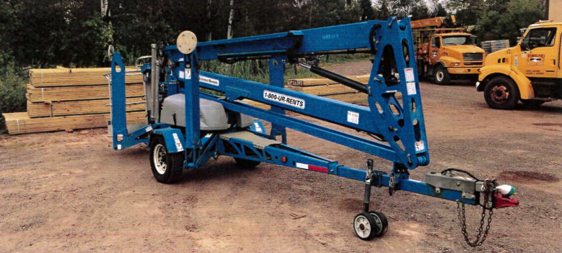 Blue Genie aerial lift on a trailer. In a gravel lot with yellow trucks and lumber.
