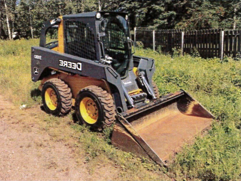 A John Deere skid steer loader with a front bucket on grassy terrain.