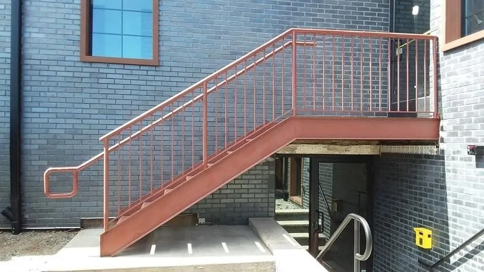 Exterior metal staircase with red railings attached to a brick building; the stairs lead down to a doorway.
