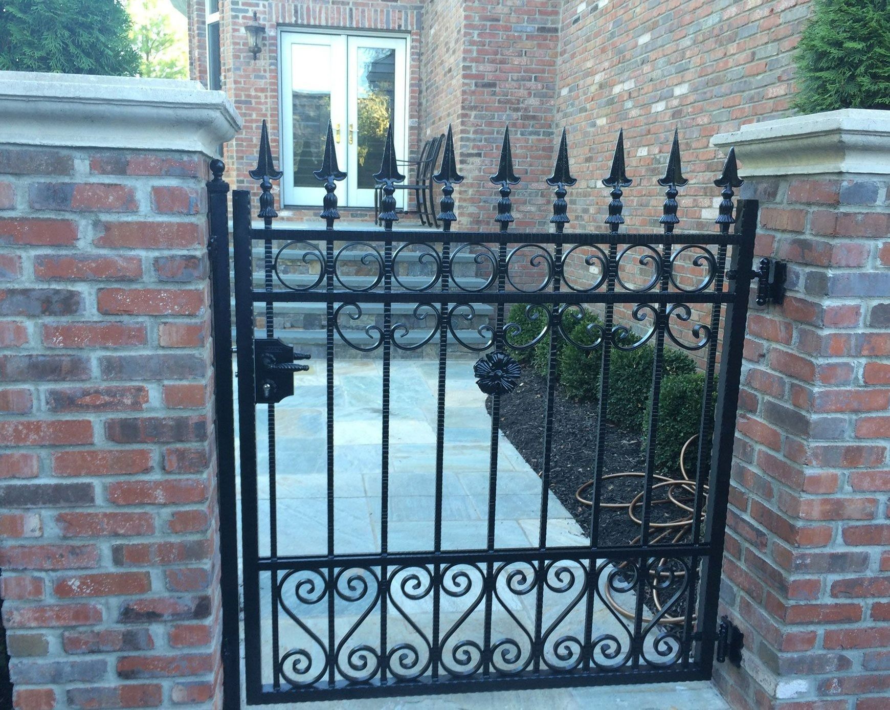 Black wrought iron gate with decorative elements set between brick pillars, leading to a path and door.