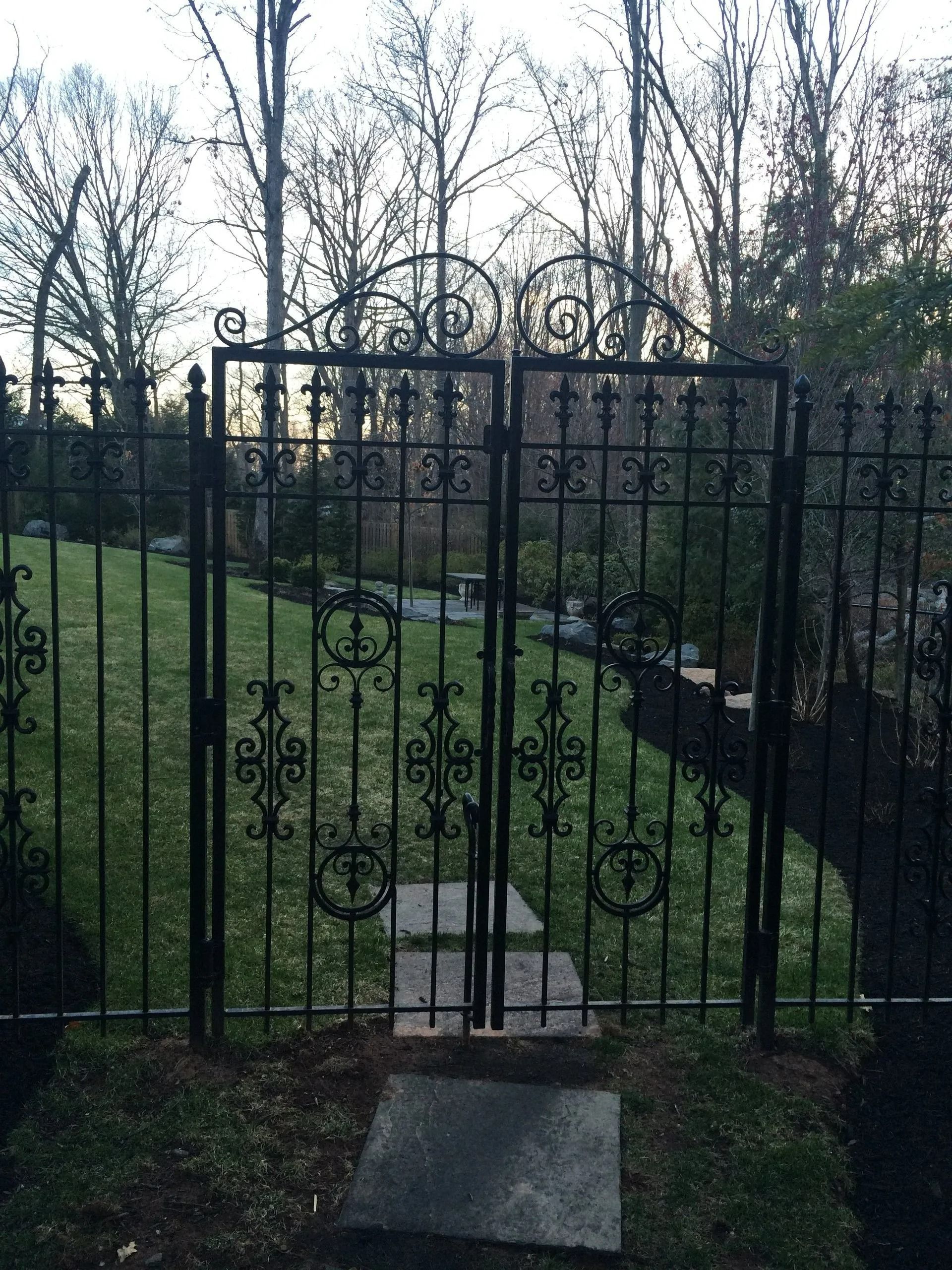 Black wrought-iron gate in a yard, leading to a stone path. Lawn and trees in background.