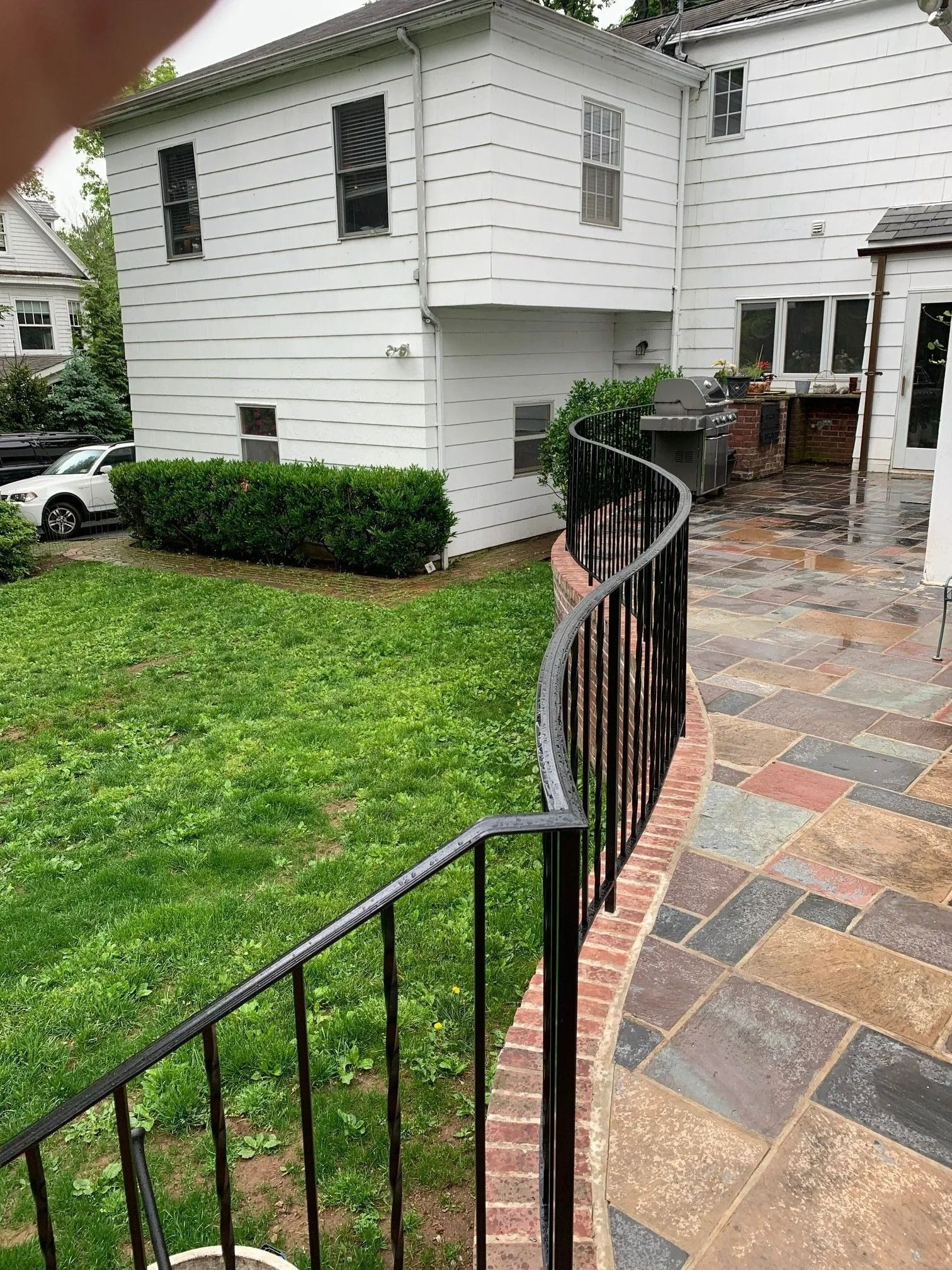 Backyard with a curved black railing, brick edging, and stone patio leading to a white house.