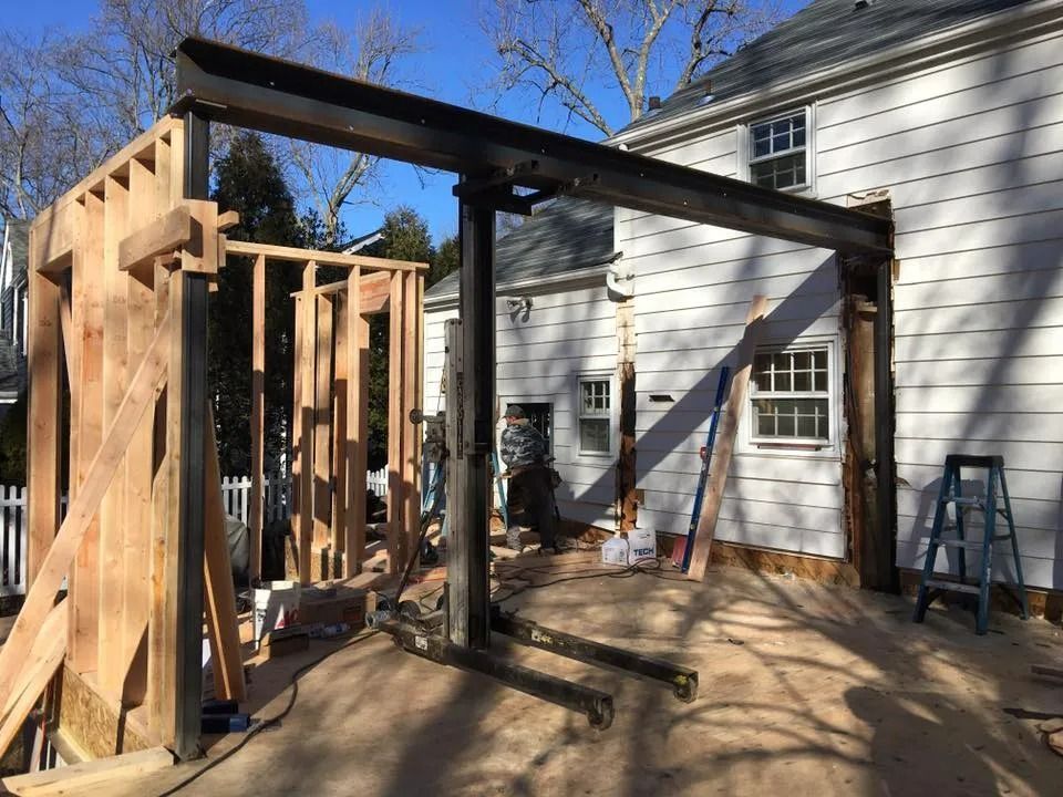 Construction site with steel beam supporting a wooden frame attached to a white house; clear sky.