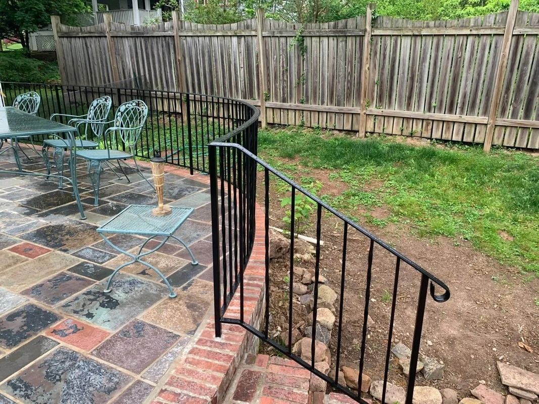 Black iron handrail on brick steps next to a patio with metal furniture, yard and wooden fence.