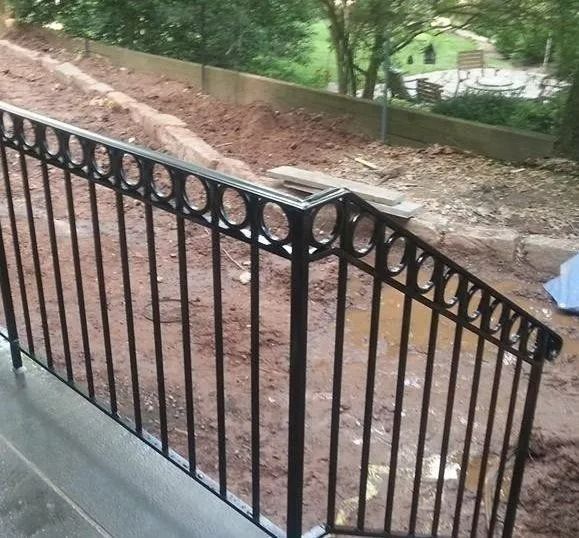 Black metal railing with circular design, outdoors. Behind, dirt slope and concrete wall.