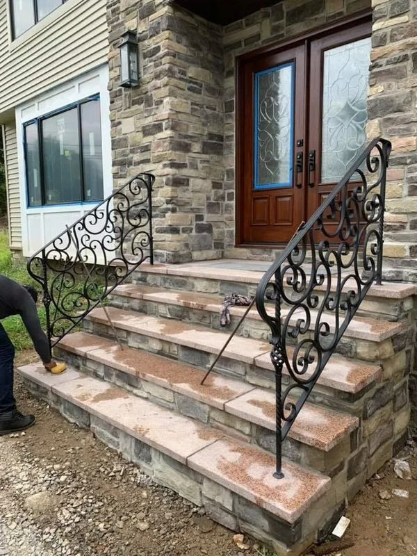 Stone steps with ornate black railing leading to a home's double doors. A person works on the steps.
