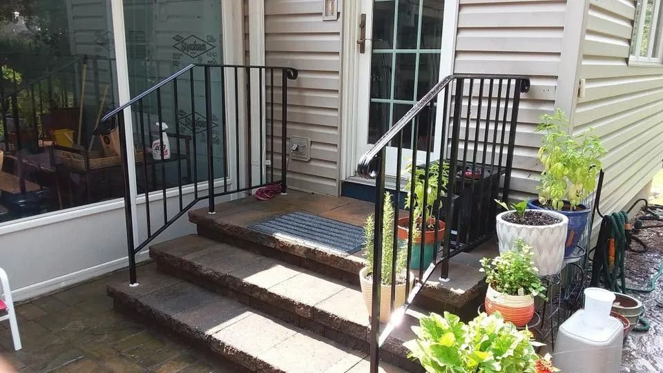 Black metal handrails on three brick steps leading to a door with plants nearby.
