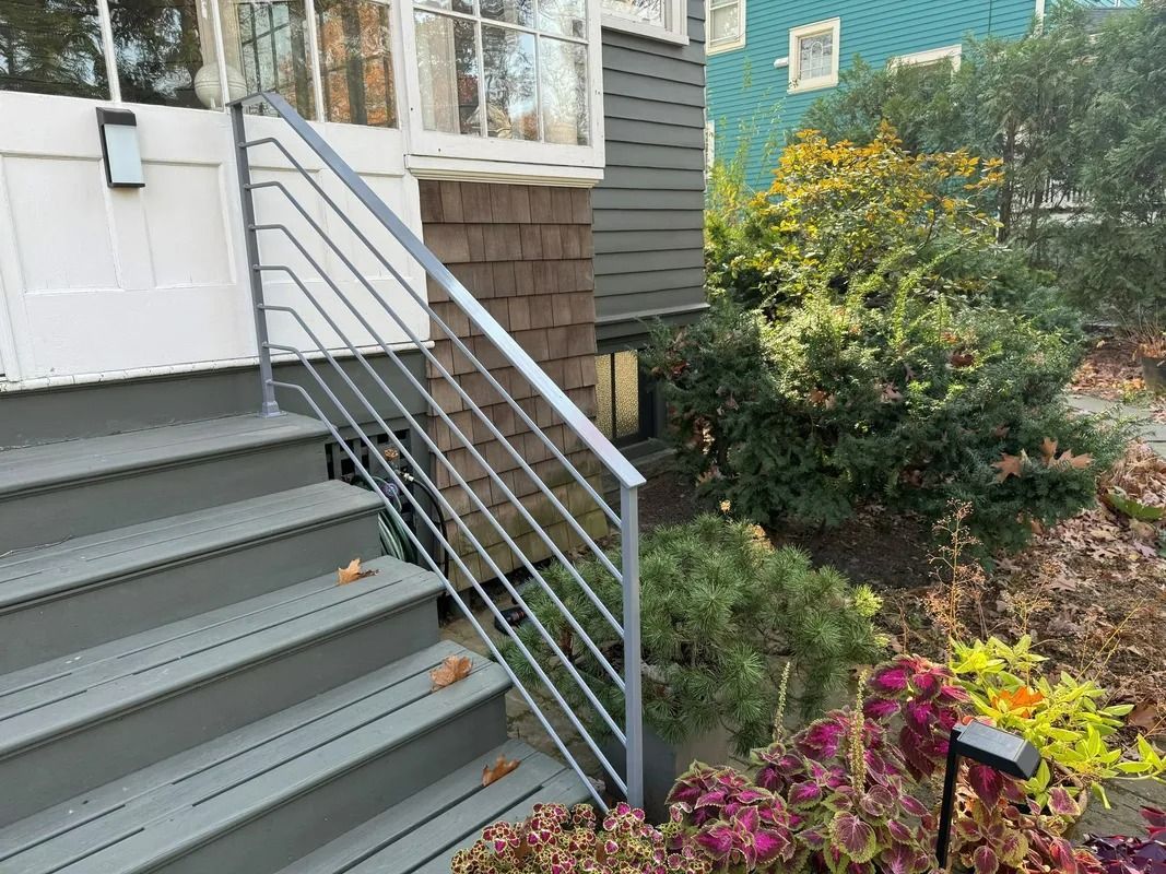 Gray stairs with a metal railing leading to a white door. A house with green and brown siding is visible.