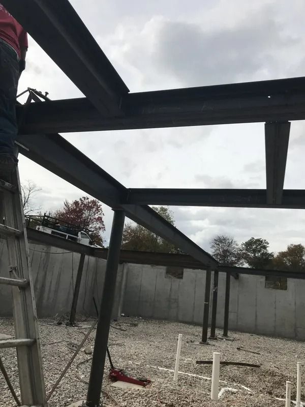 Construction of a steel frame structure. A person stands on a ladder, working on the beams. Gray sky and gravel ground.