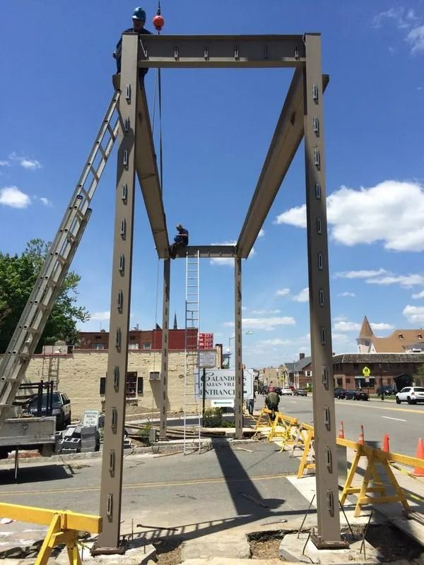 Steel framework being constructed over a street, two workers, ladder, blue sky, and surrounding buildings.