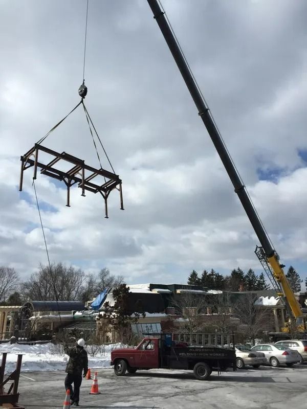 A crane lifts a metal structure at a construction site. A worker watches nearby, a red truck is parked.