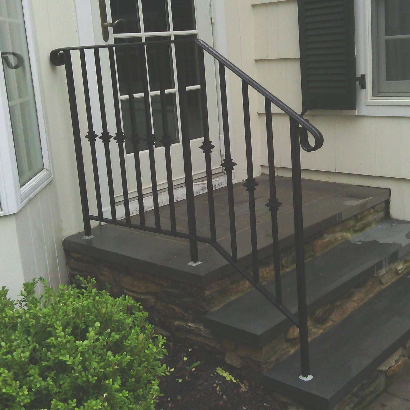 Front steps with black metal railing leading to a porch beside light-colored siding and a green shrub.