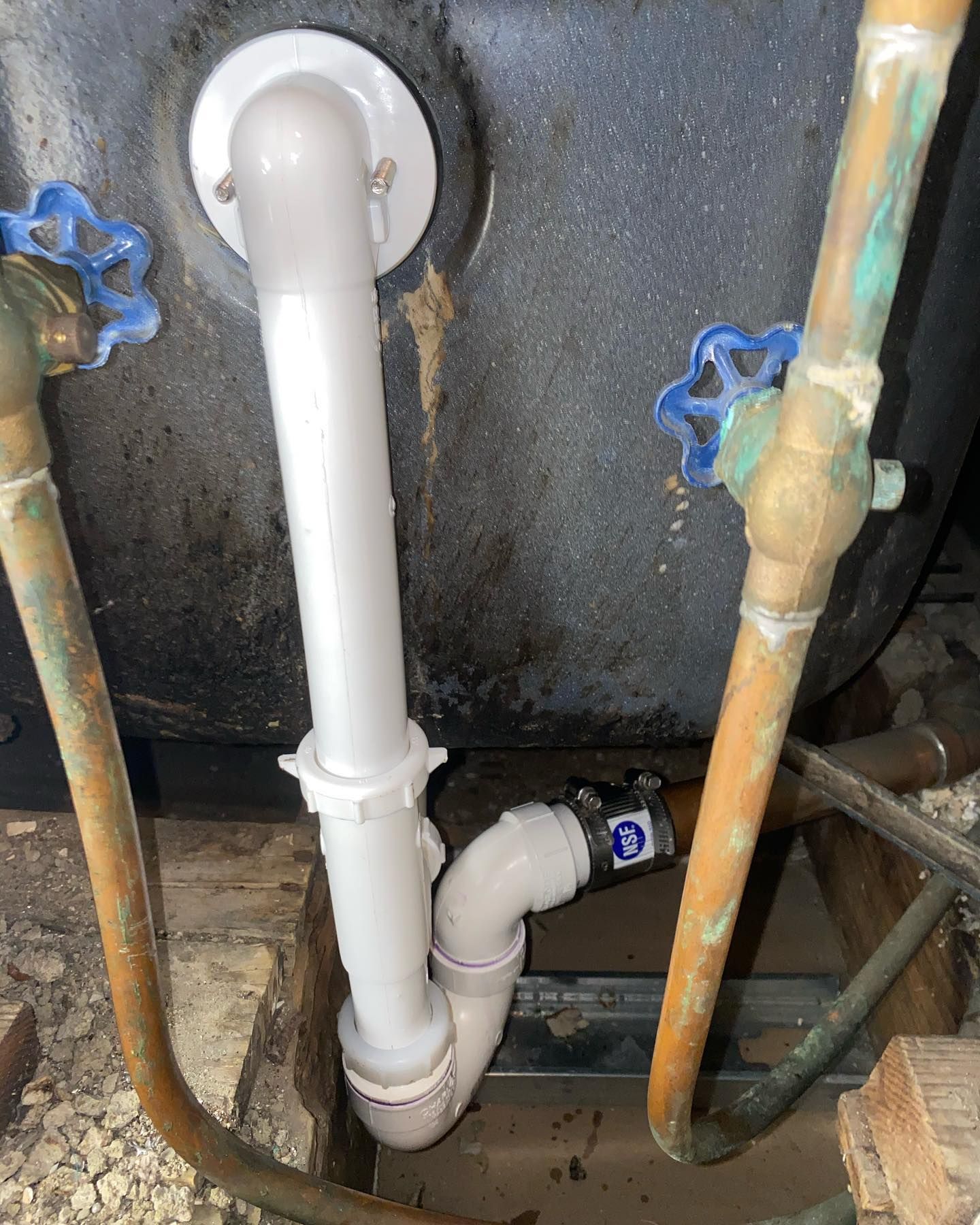 Plumbing of bathtub: white drainpipe, water pipes with blue handles, black tub, and wood framework.