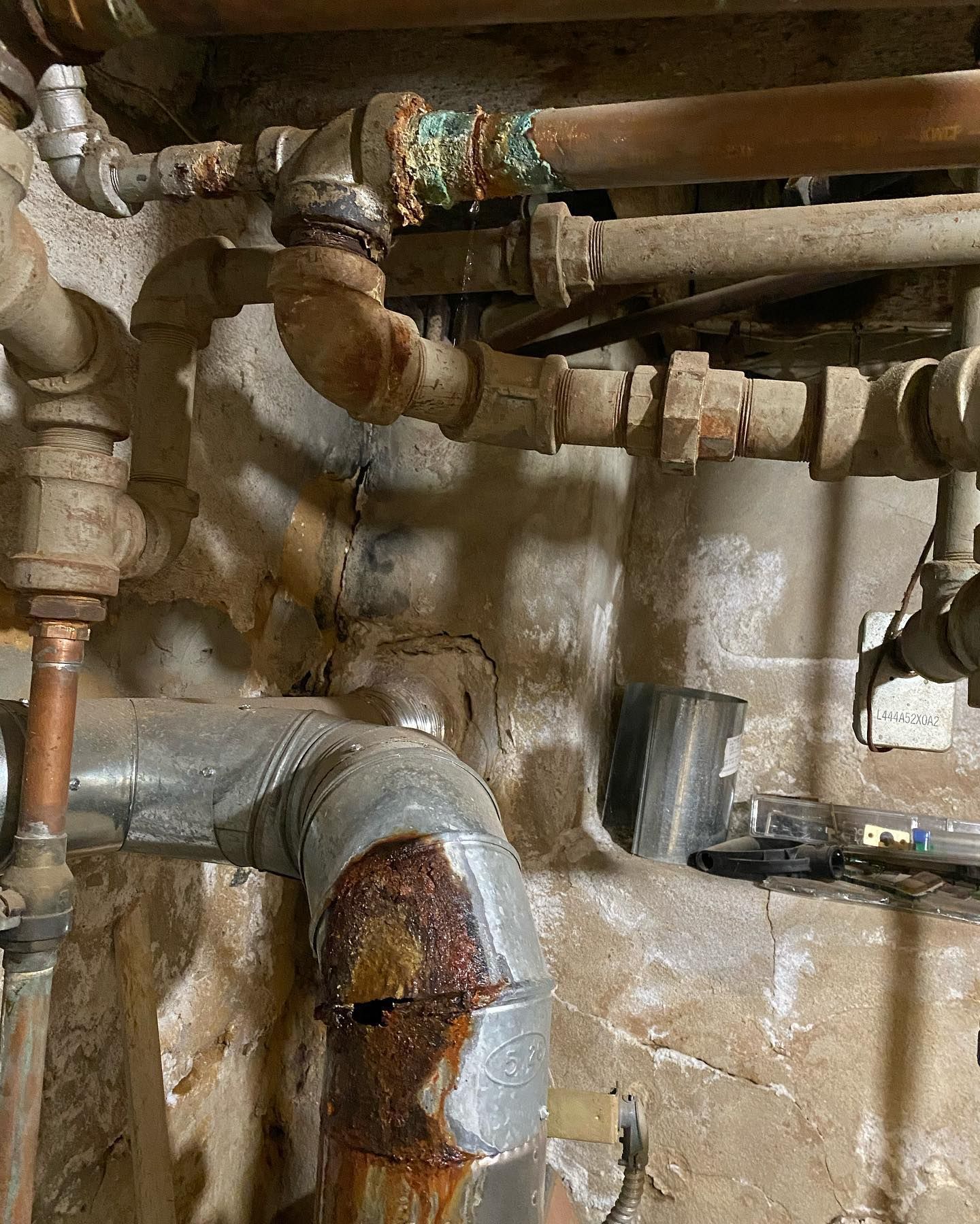 Rusty pipes and fittings in a dimly lit basement. Visible water damage on the wall.