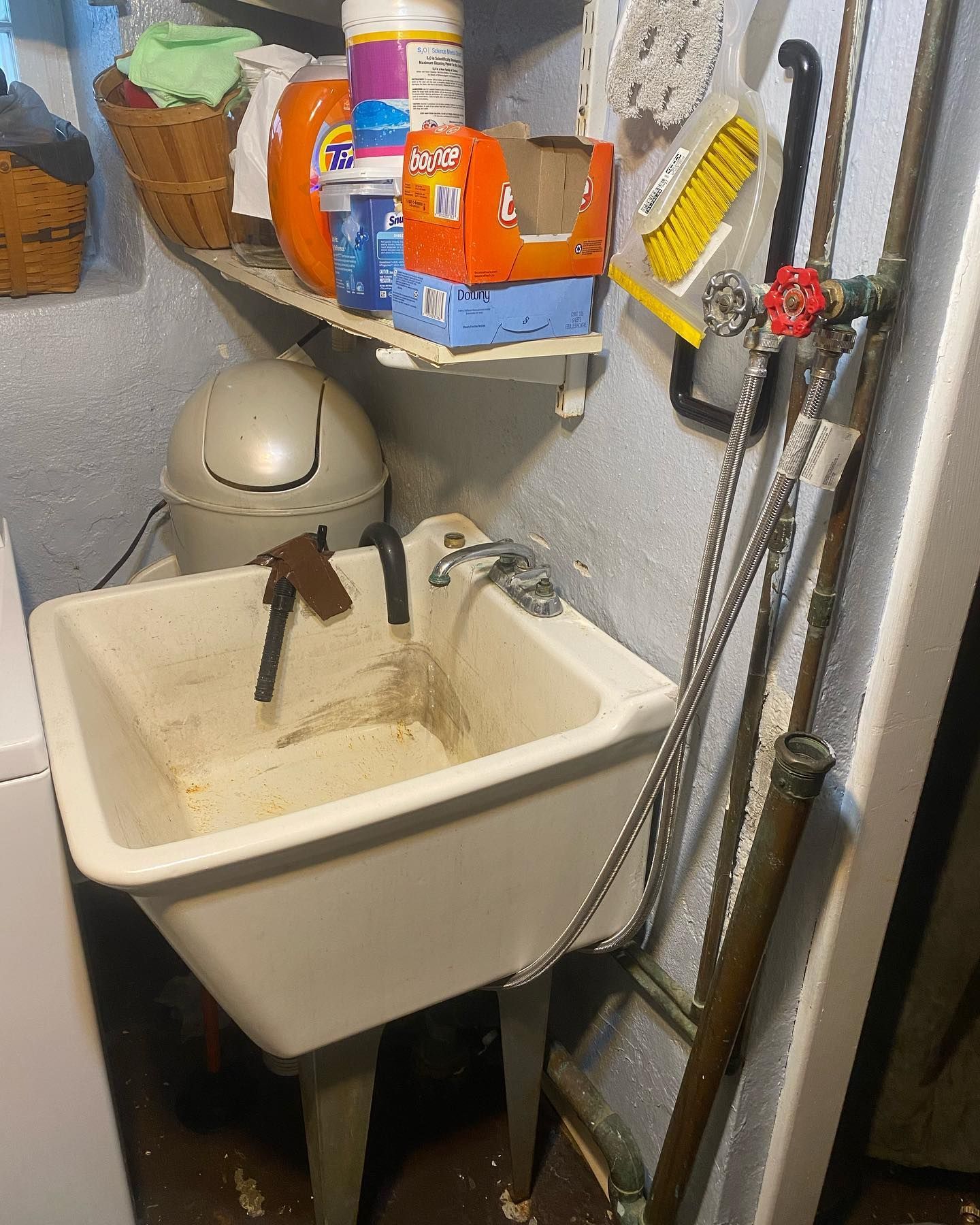 Laundry sink in cluttered utility room with cleaning supplies and trash can.