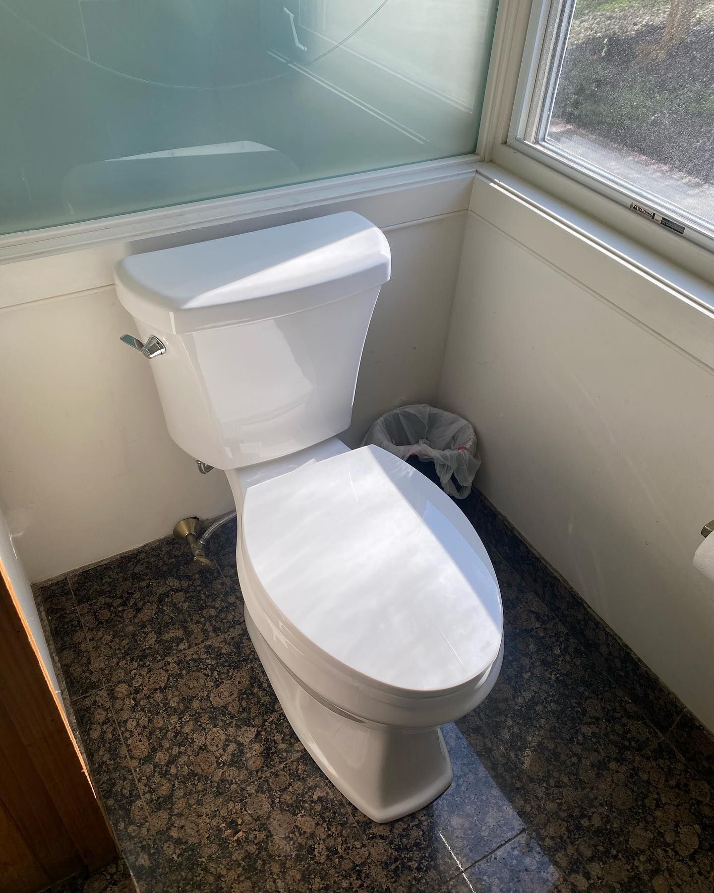 A white toilet in a small bathroom with a window and a trash can; granite floor.