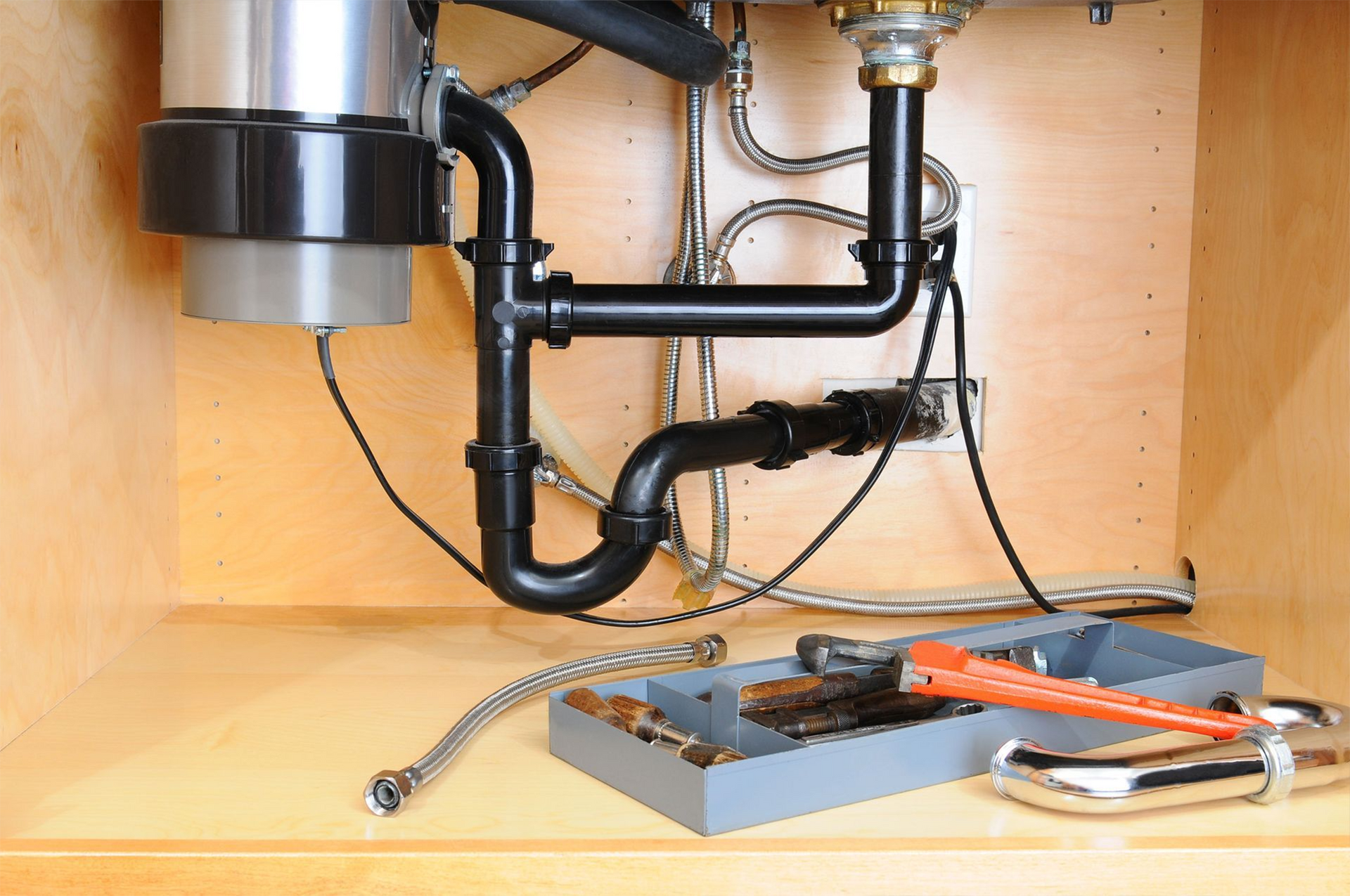 Garbage disposal and plumbing under kitchen sink, with tools on the shelf.