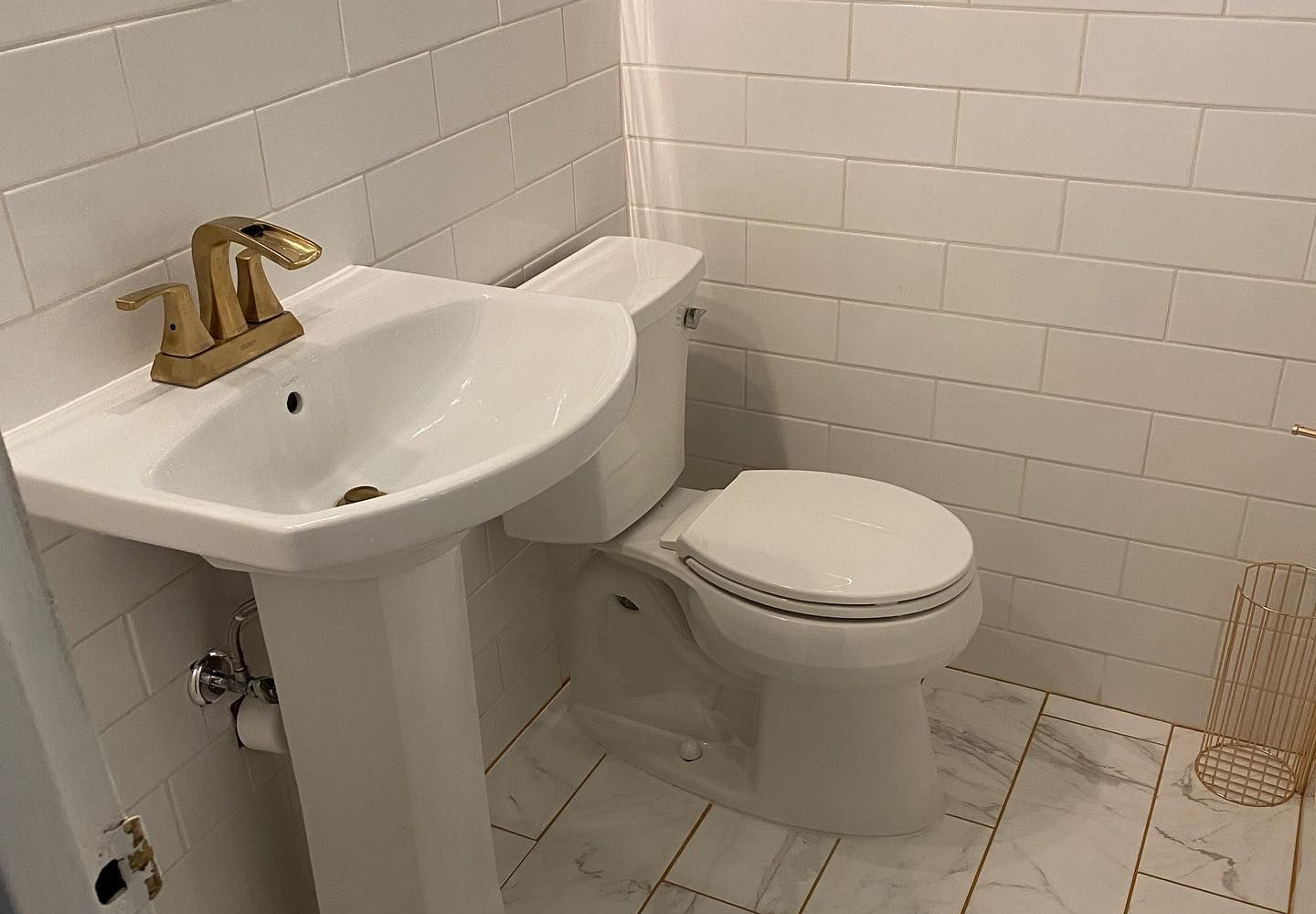 White bathroom with pedestal sink, gold faucet, and toilet against white subway tile.