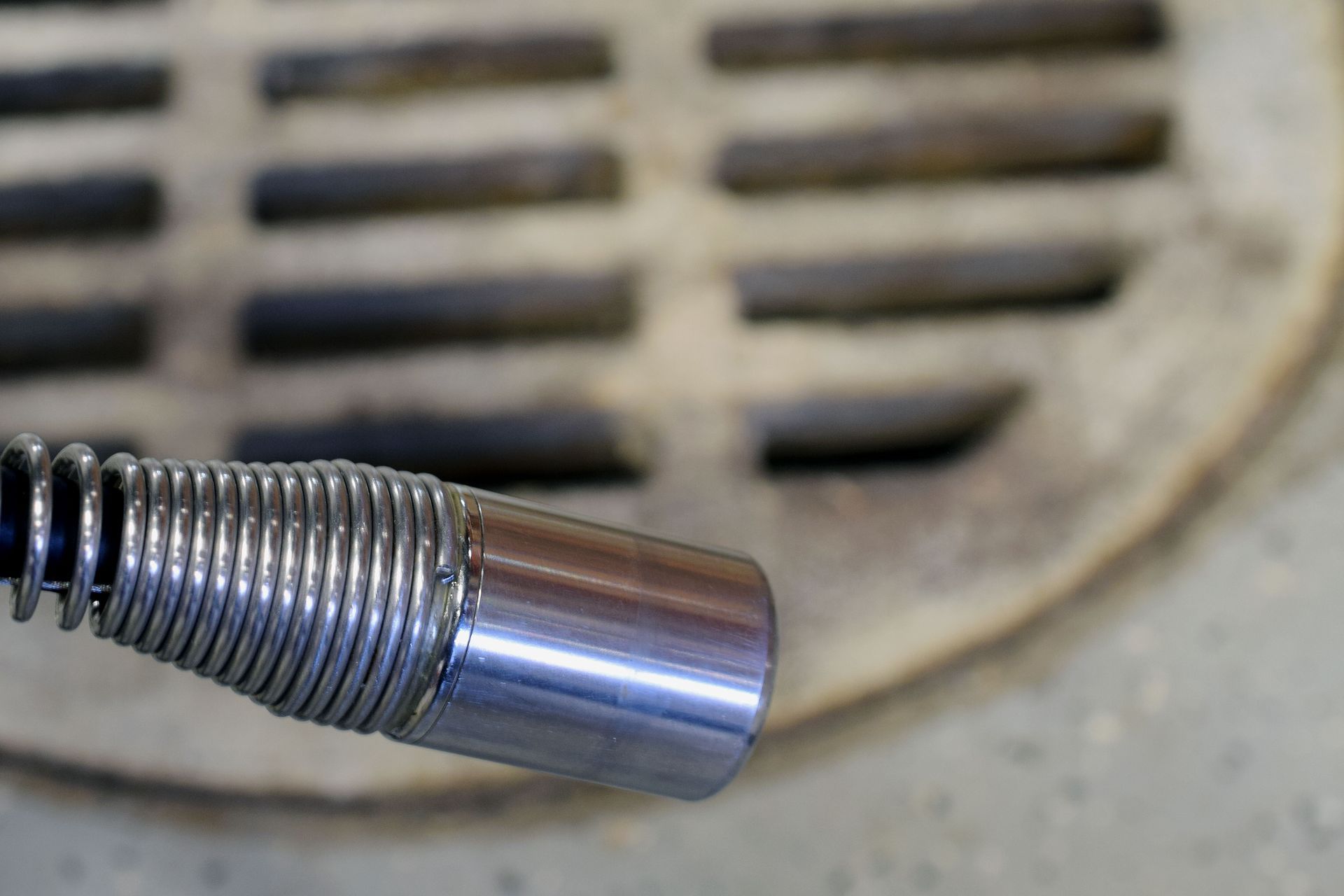 Drain snake head inserted into a round, slotted drain. Shiny metal against a dull, off-white surface.