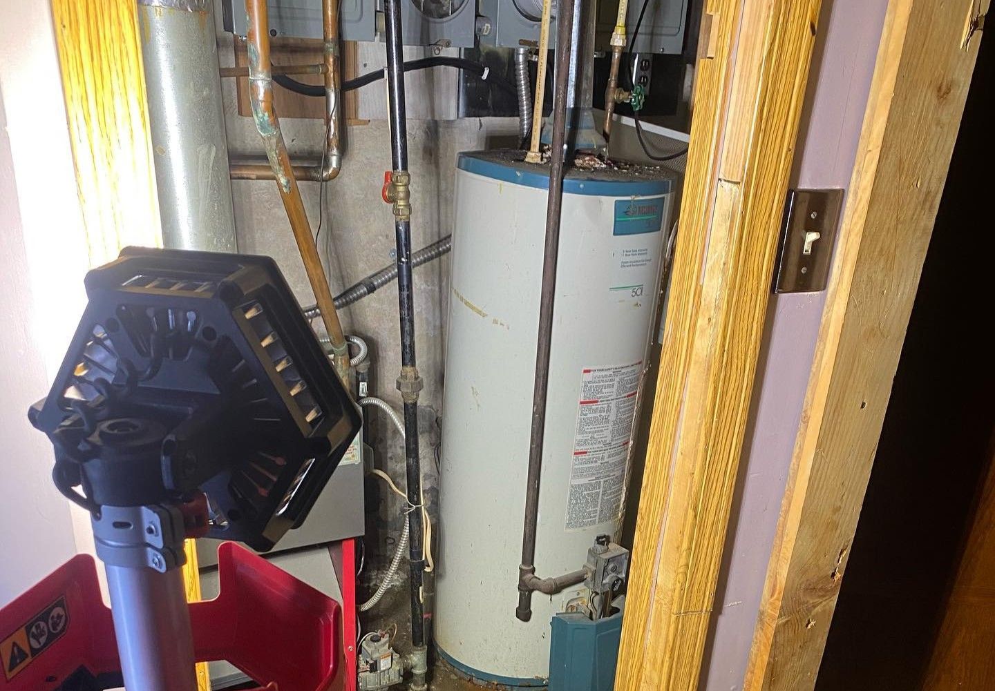 Water heater in a utility room, surrounded by pipes. A work light is in the foreground.