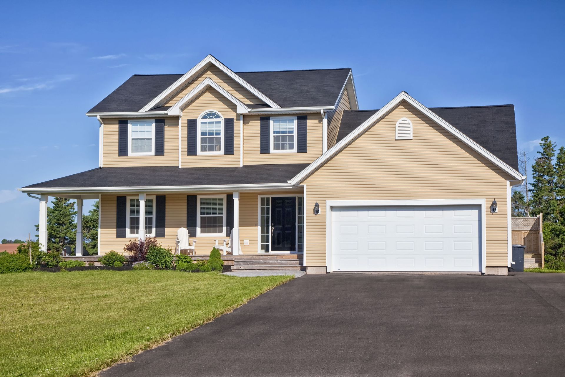 Tan two-story house with black roof and garage. Black asphalt driveway, green lawn, and blue sky.