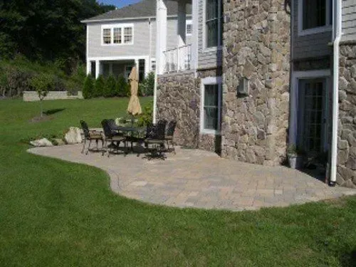 Stone patio with outdoor dining table, adjacent to a house with stone facade, on a green lawn.