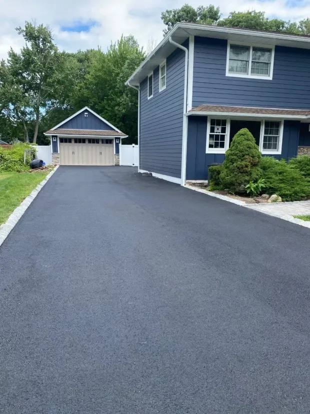 Long black paved driveway leading to a blue two-story house and a detached garage, all under a cloudy sky.