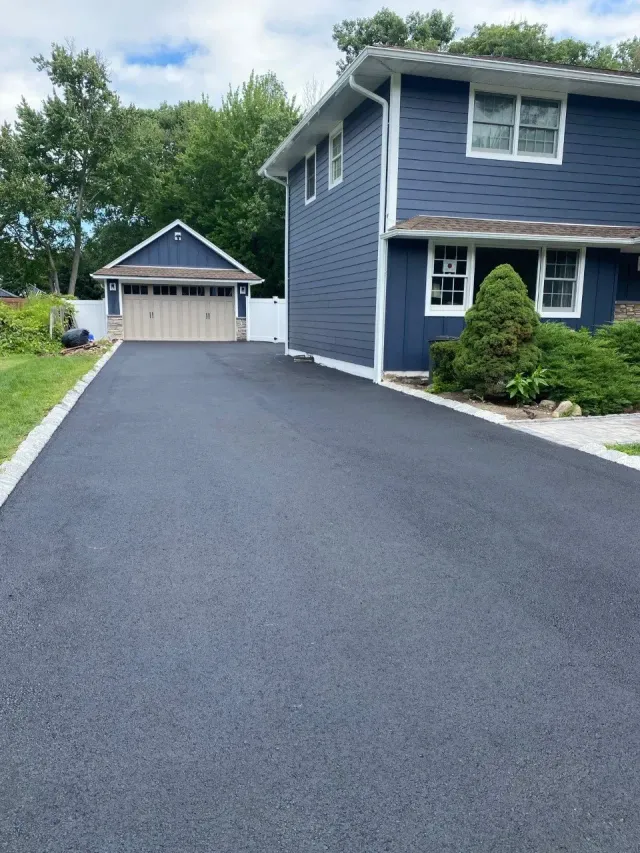 Long black paved driveway leading to a blue two-story house and a detached garage, all under a cloudy sky.