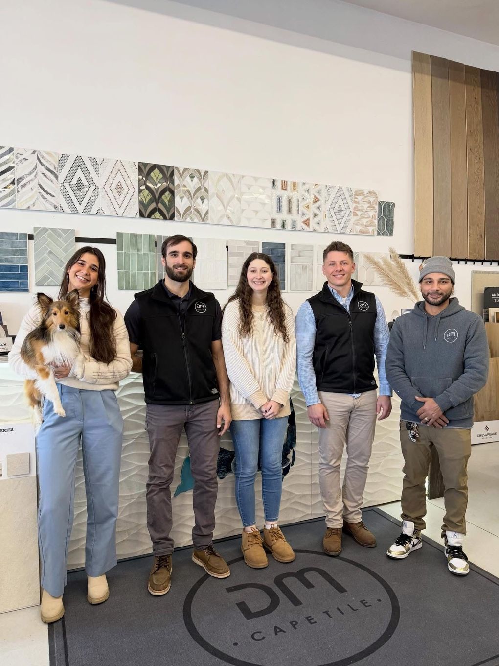 Five people standing in a showroom with tile samples. A woman holds a small dog. 