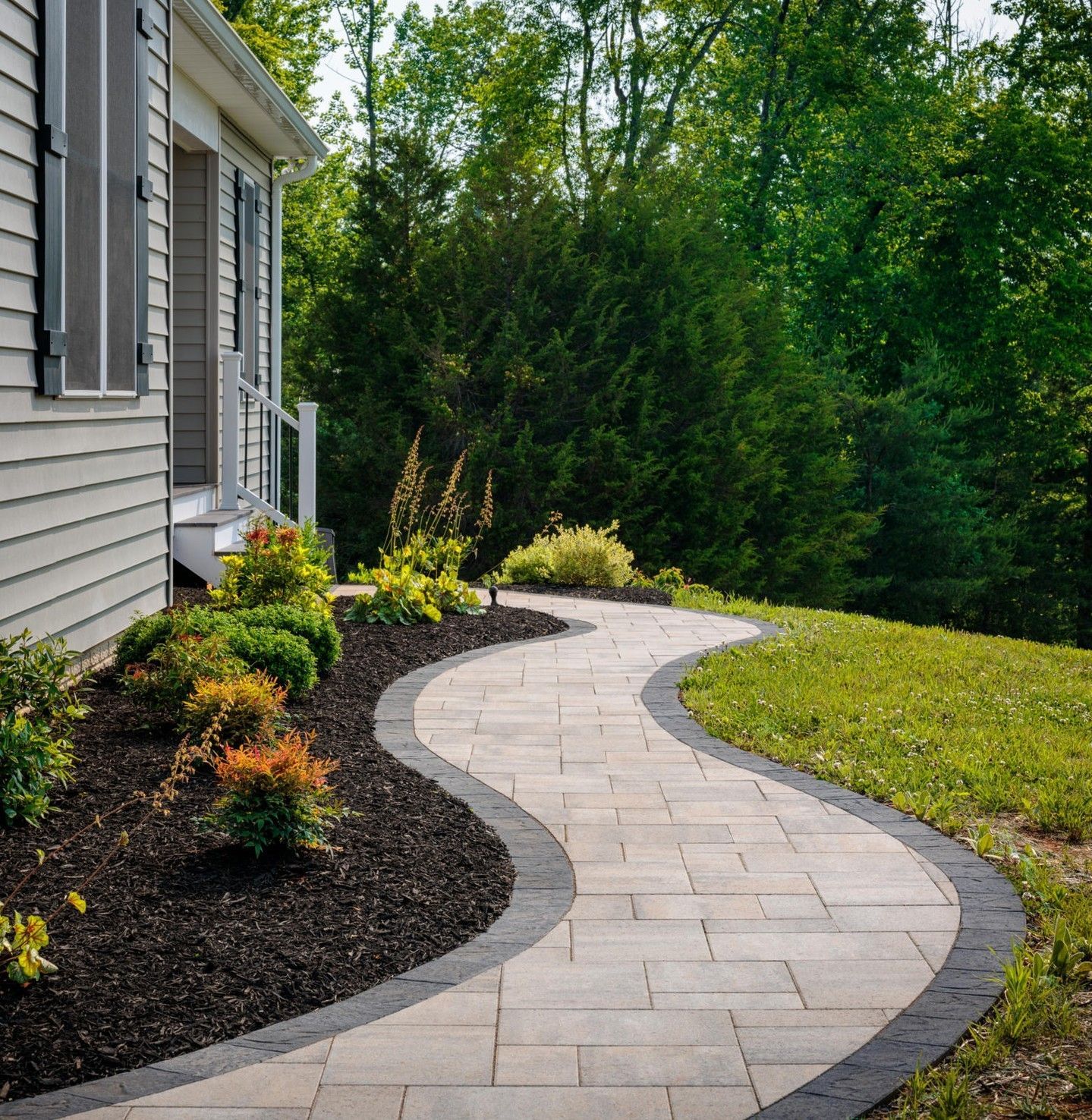 Curving brick walkway bordered by dark edging, leading through garden to a house.