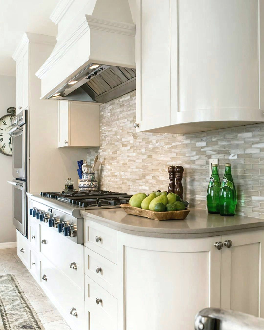 White kitchen with range, tiled backsplash, and curved countertop. Green bottles and fruit add color.