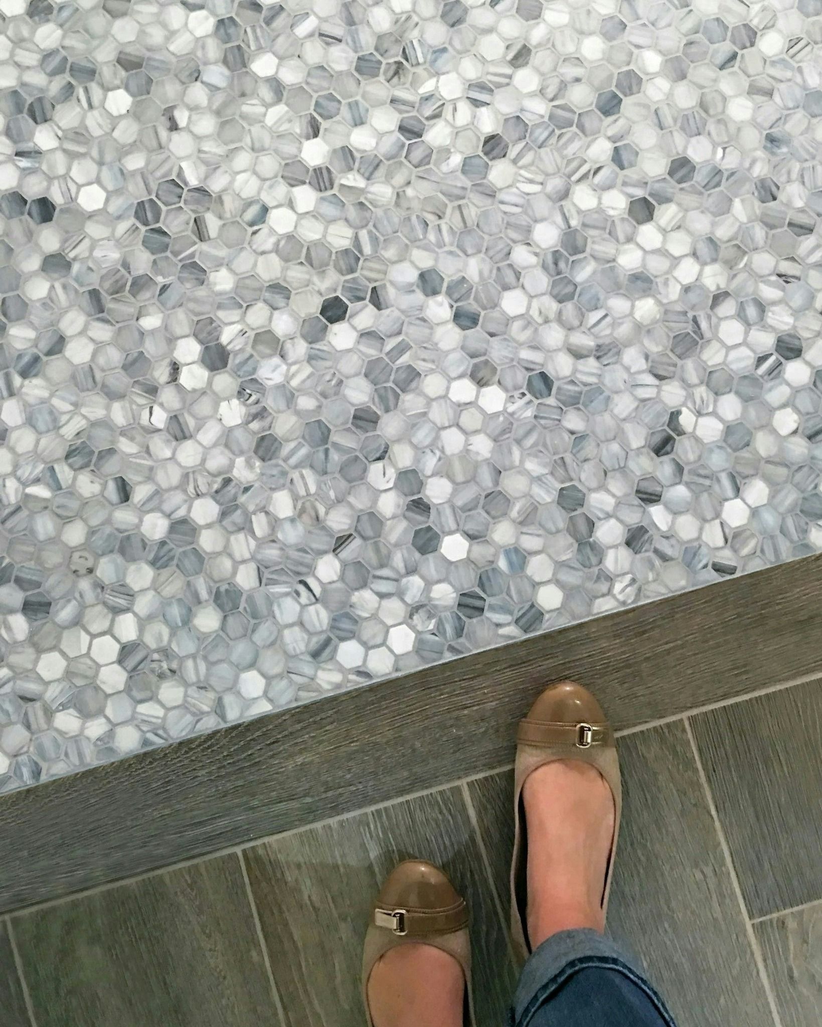 Person's feet in tan flats stand on gray tile, next to patterned mosaic tile floor, viewed from above.