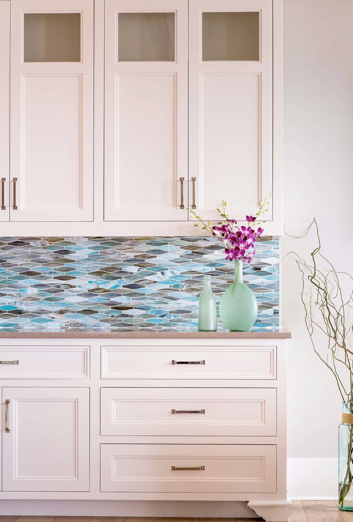 White kitchen cabinets with blue mosaic backsplash, purple flowers in green vase.