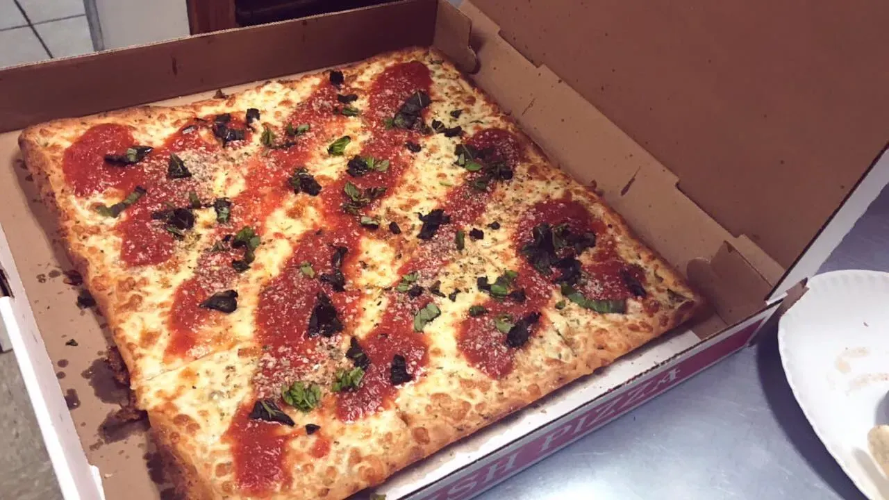 Square pizza in a cardboard box, topped with sauce and basil.