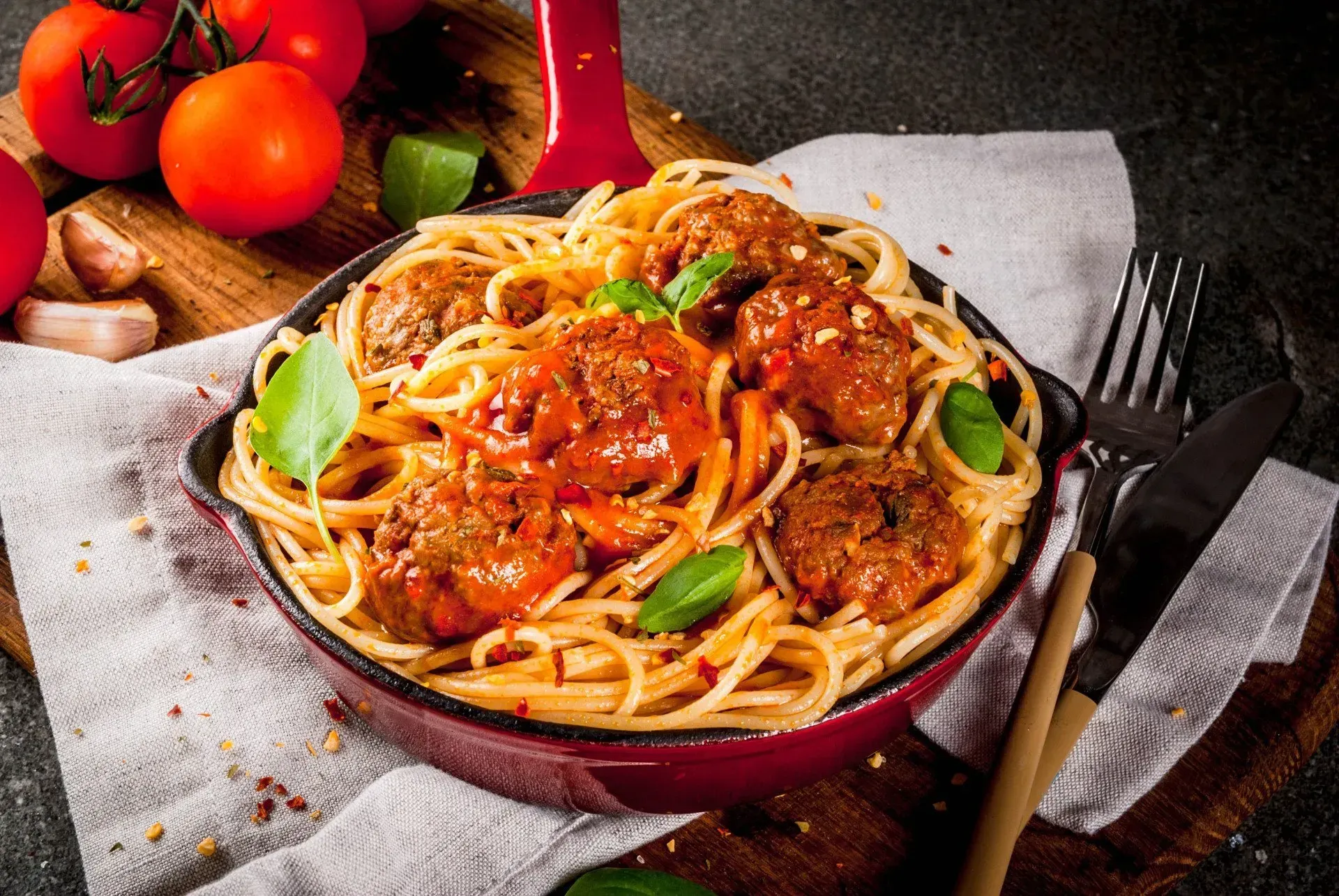 Spaghetti and meatballs in a red pan, with tomatoes, basil, and a knife and fork on a wooden board.