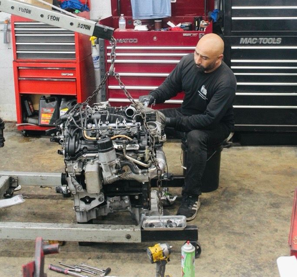Mechanic in red shirt and overalls working on a car engine in a garage.