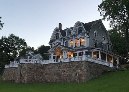 Large gray house on a stone retaining wall, with a wraparound porch, and a lawn.