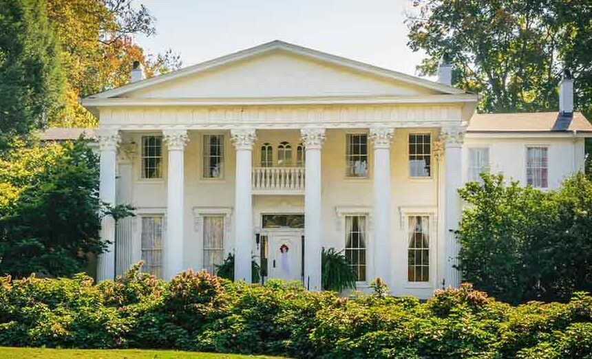 White Greek Revival-style mansion with tall columns and lush greenery in front.