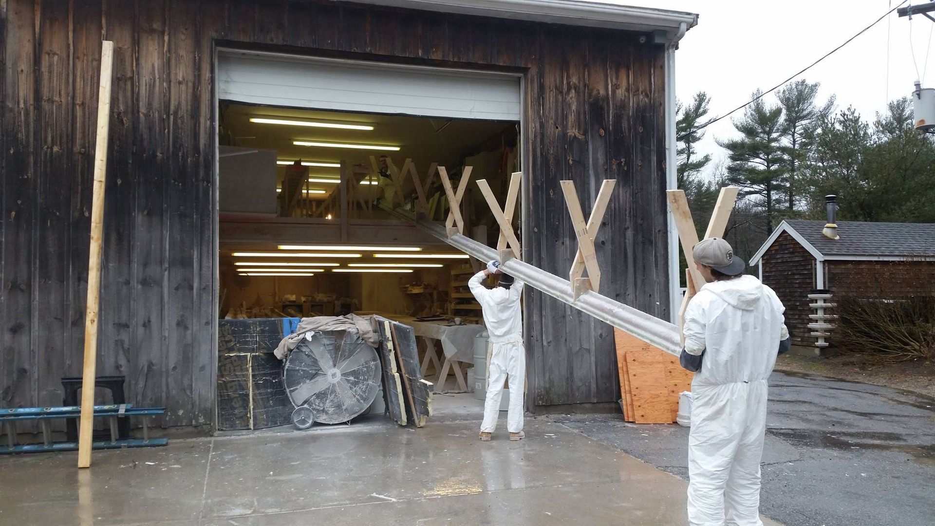 Two people in white coveralls carrying a long metal beam outside a wooden barn.