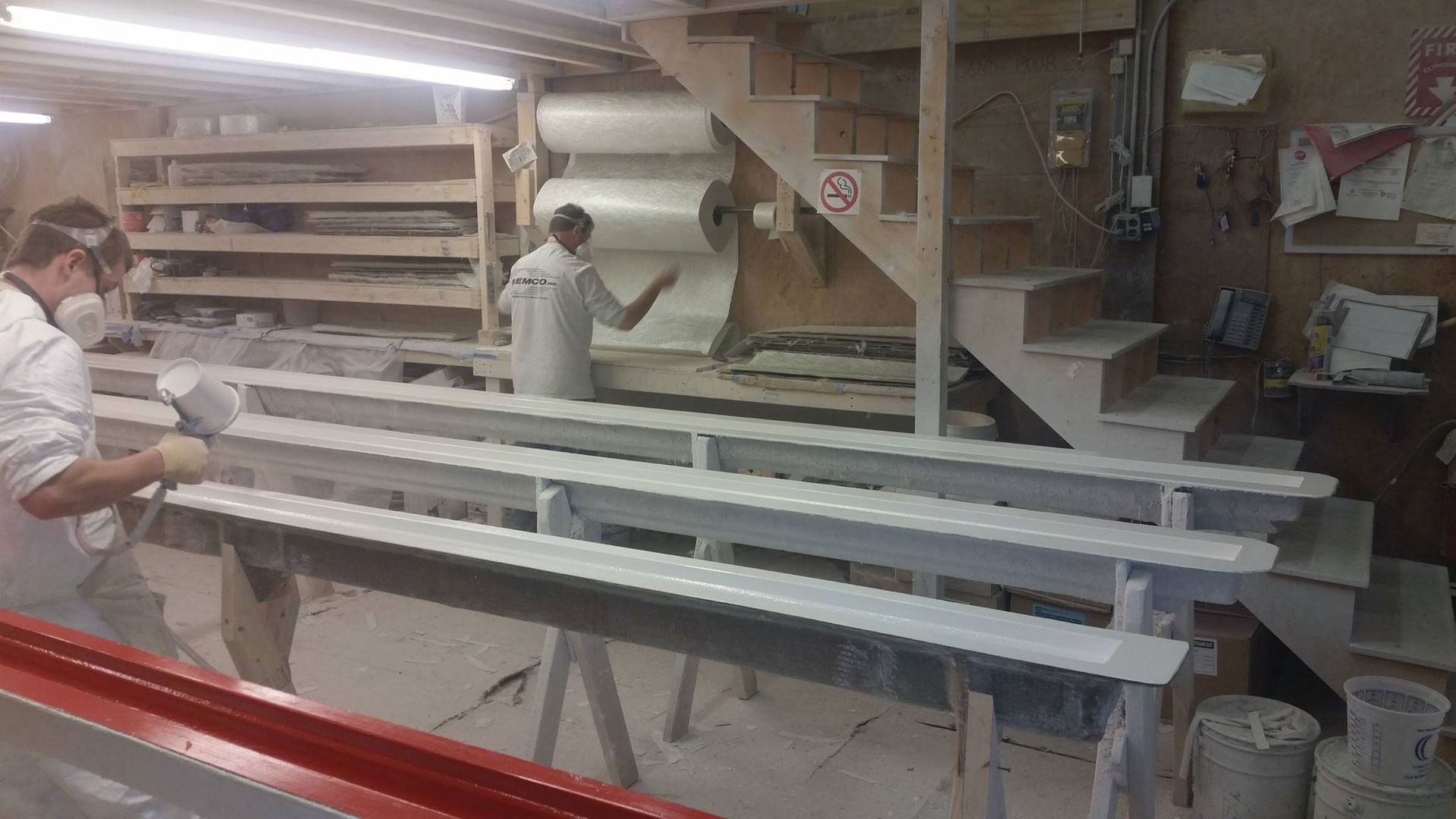 Man spraying white paint on beams in a workshop, another worker in background.