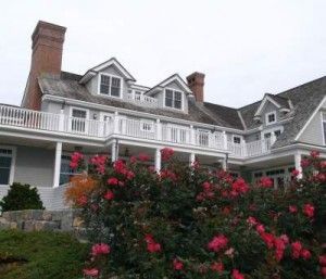 Gray house with white trim, red brick chimneys, and a second-story balcony, in front of pink rose bushes.