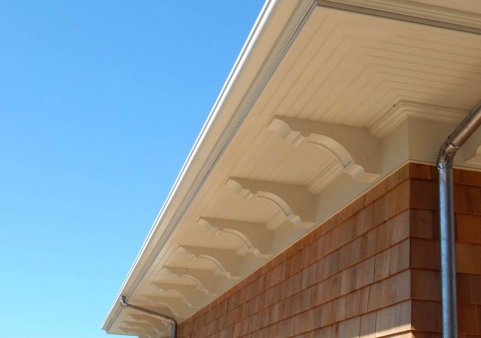 Beige roof overhang with decorative brackets over a brown shingled wall against a blue sky.