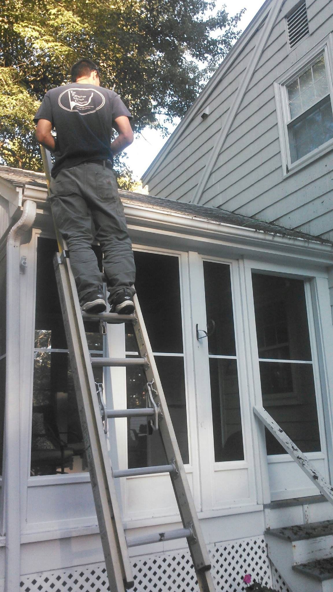 Person on ladder cleaning gutters of a white house with a porch.