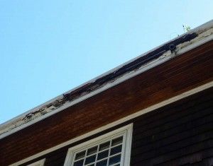 Damaged roofline with dark wood siding, white window frame, and clear blue sky.