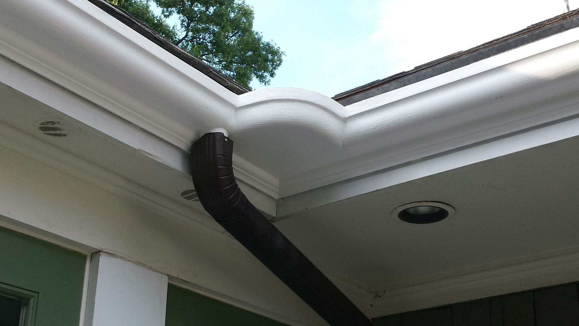 White guttering on a house with a black downspout. Sky and trees in the background.